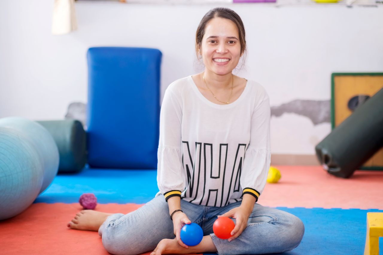 about-01 Smiling woman holding therapy balls, seated in a brightly colored fitness studio.