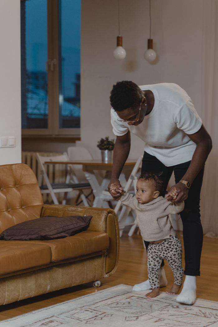 gallery-2 A father assists his toddler in walking inside a cozy living room.
