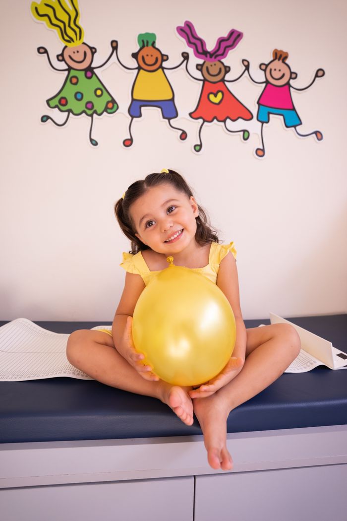 hero-img-02 Happy child sitting on medical table holding yellow balloon, smiling.